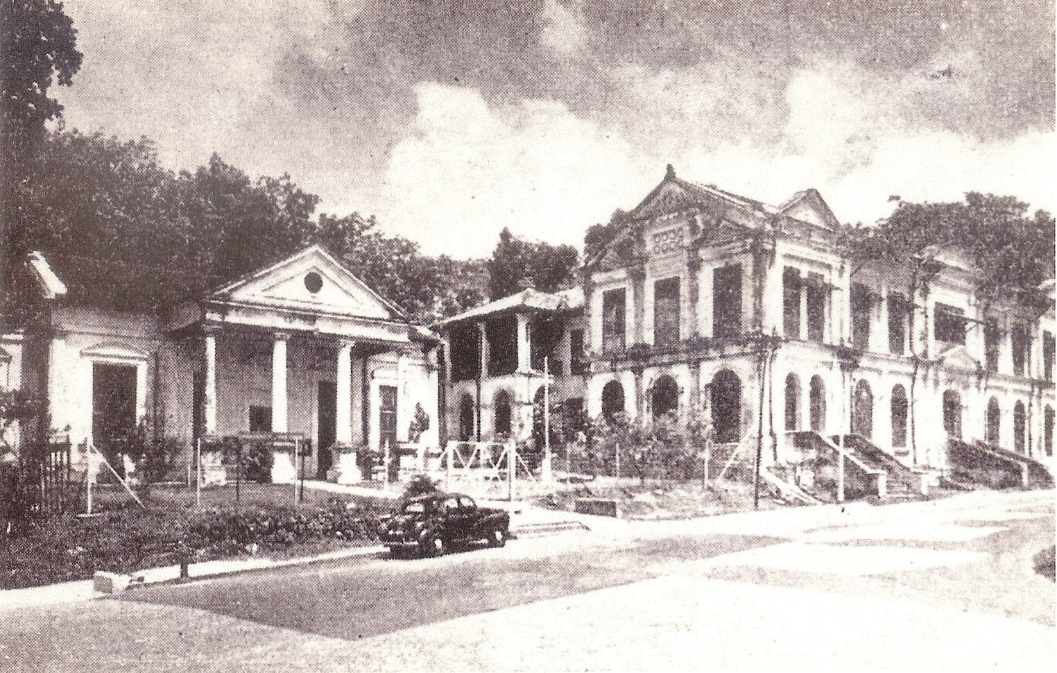 Old black and white photo of two colonial-style buildings with columns, a vintage car parked in front, and trees behind.